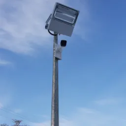 Słup oświetleniowy z kamerą przemysłową na tle błękitnego nieba, widoczne budynki w tle pokryte śniegiem.