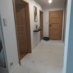 Zbigniew Z. - Nowoczesny korytarz z białymi płytkami imitującymi marmur, drewnianymi drzwiami i listwami przypodłogowymi, oraz minimalistyczną półką z dekoracjami.