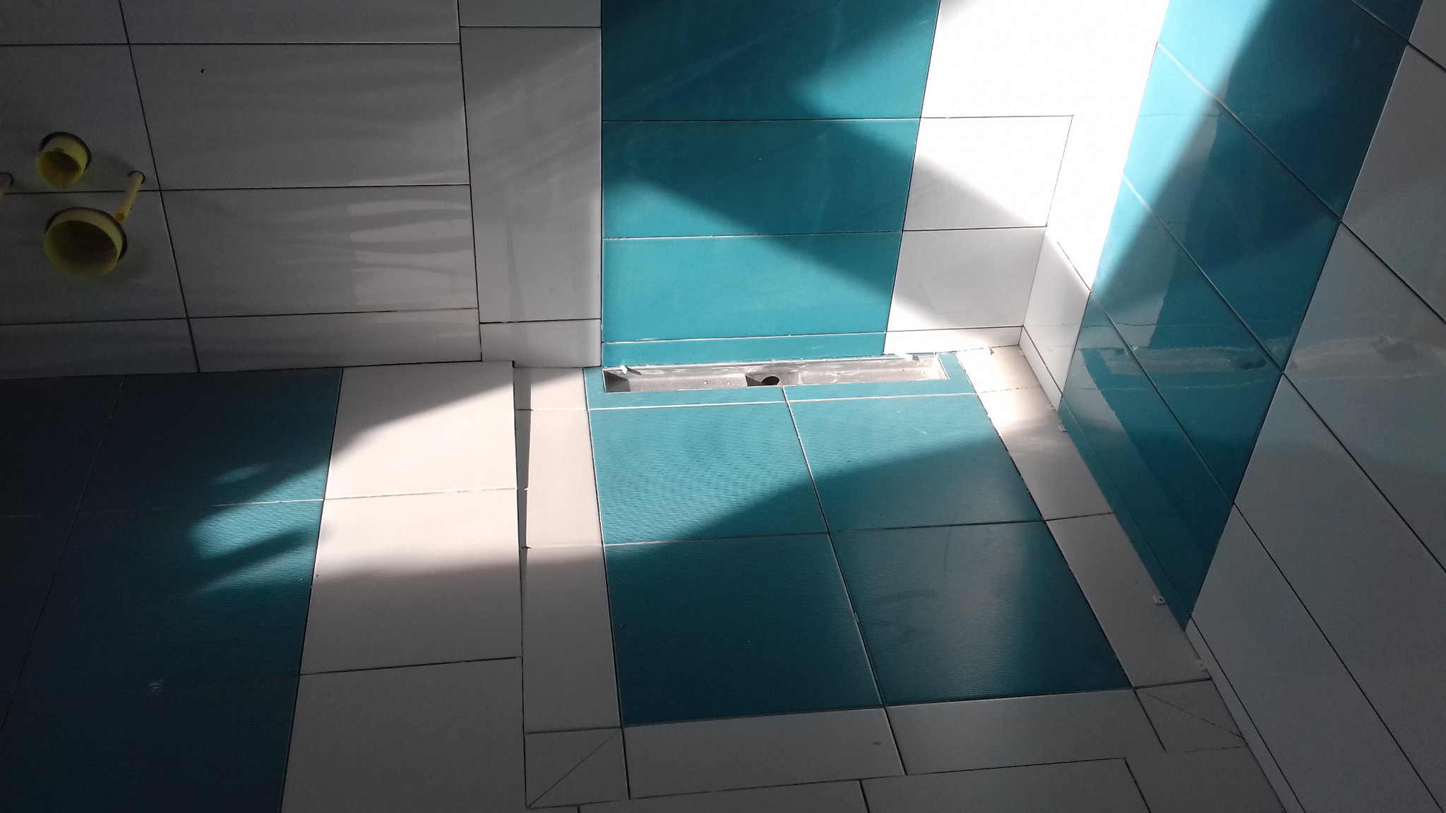 Nowoczesna łazienka w trakcie wykańczania, widoczne białe i turkusowe płytki, odpływ liniowy w podłodze i wystające rury instalacji wodnej.