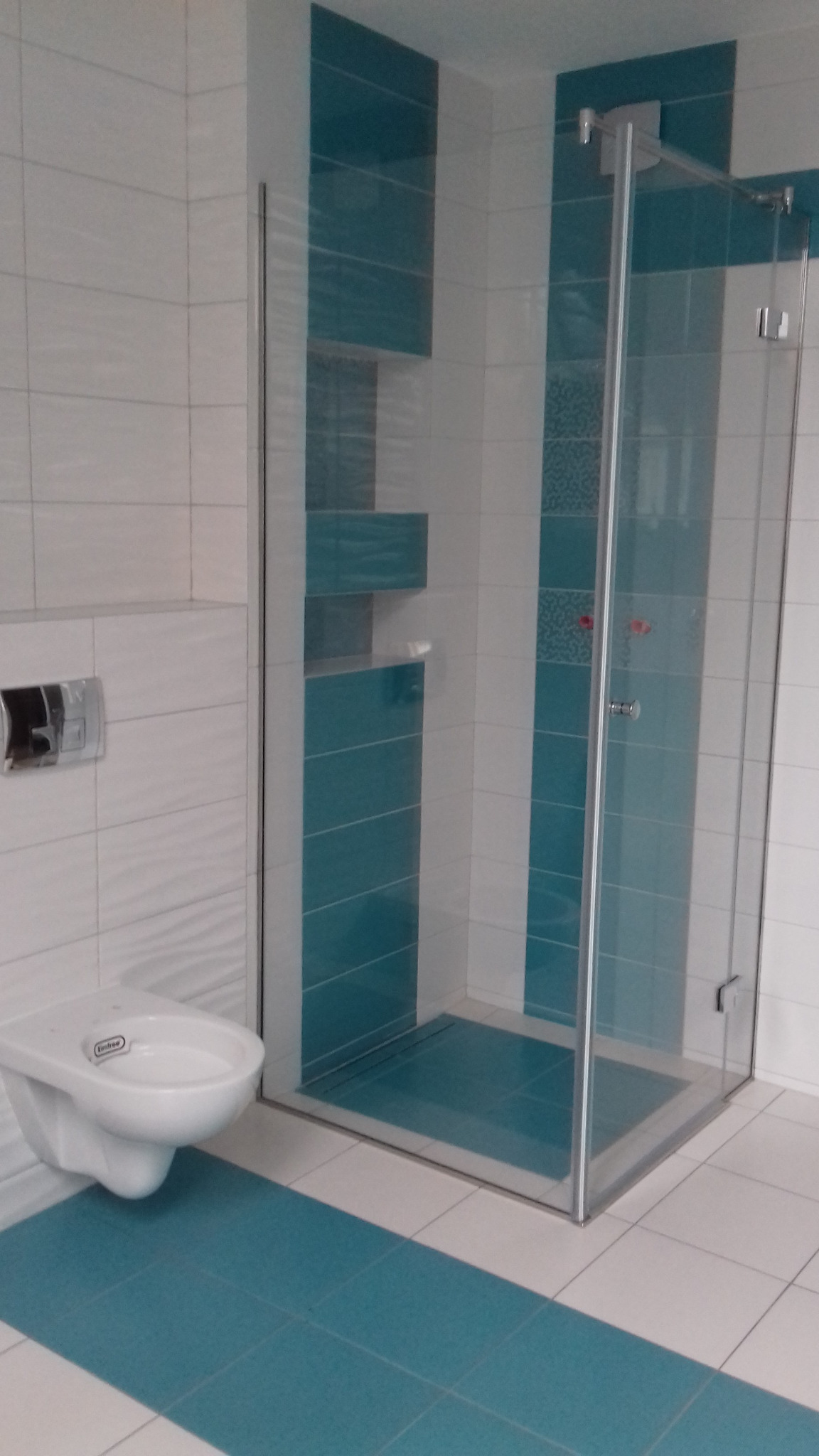 Nowoczesna łazienka z kabiną prysznicową typu walk-in, biało-turkusowe płytki ułożone w pasy, podwieszana toaleta z chromowanym przyciskiem spłukującym.