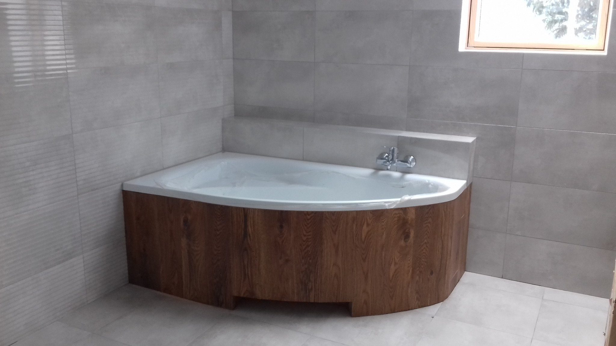 Narożna wanna obudowana panelami z imitacją drewna w łazience z szarymi płytkami, widoczny kran i okno.