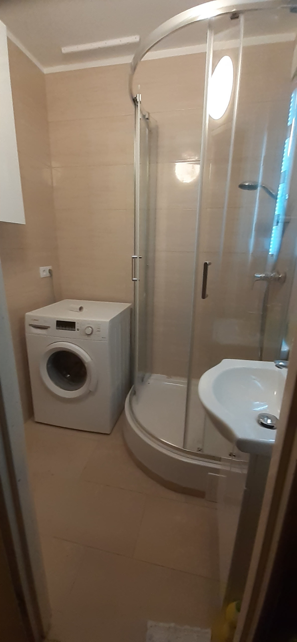 Niewielka łazienka z kabiną prysznicową z zaokrąglonymi drzwiami, białą umywalką na postumencie i pralką Bosch. Beżowe płytki ścienne i podłogowe.