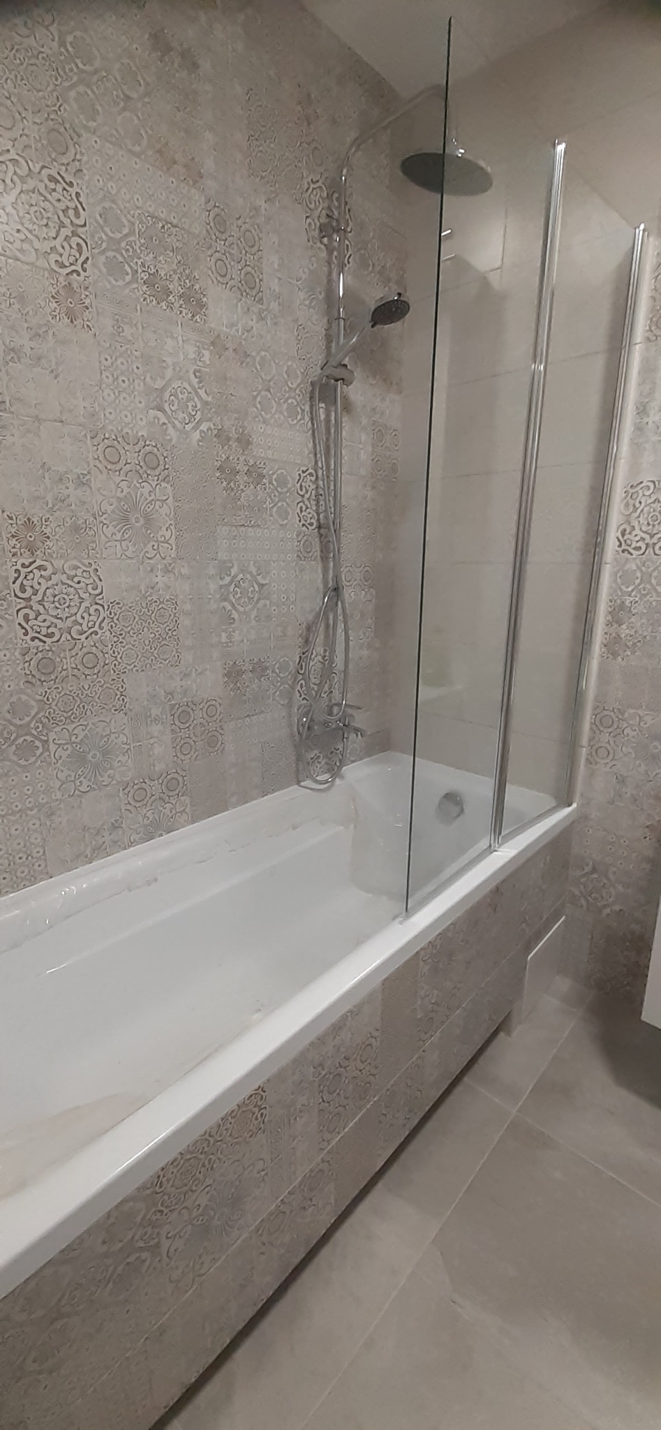 Biała wanna z zamontowaną szklaną kabiną prysznicową, ściany i obudowa wanny wyłożone płytkami w odcieniach szarości z subtelnym wzorem, widoczny zestaw prysznicowy.