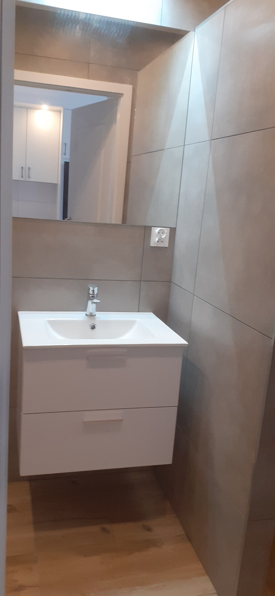 Nowoczesna łazienka z umywalką nablatową na białej szafce z dwiema szufladami, szare płytki na ścianach i lustro z widocznym odbiciem fragmentu innego pomieszczenia.