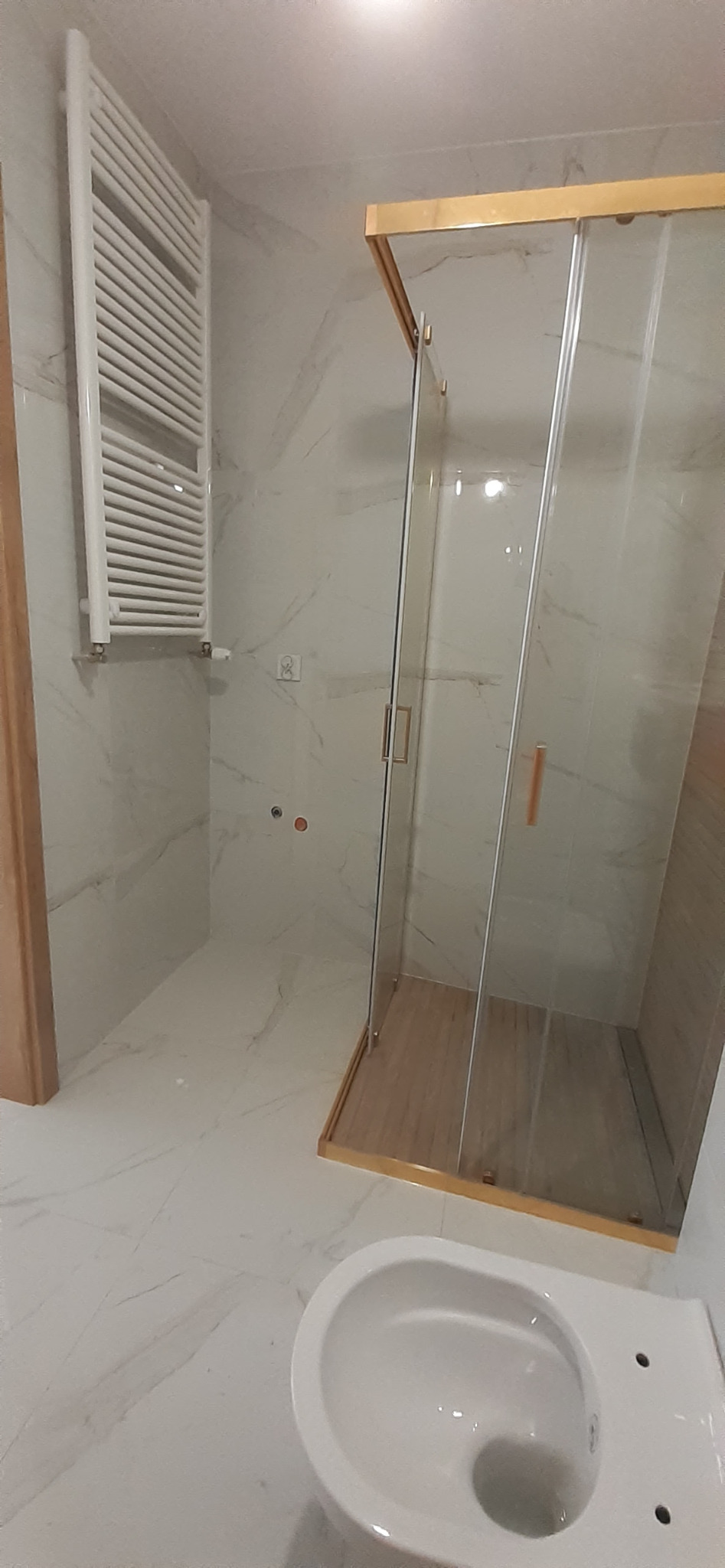 Nowoczesna łazienka po remoncie: kabina prysznicowa ze złotymi ramami, białe marmurowe płytki, biały grzejnik drabinkowy i podwieszana toaleta.