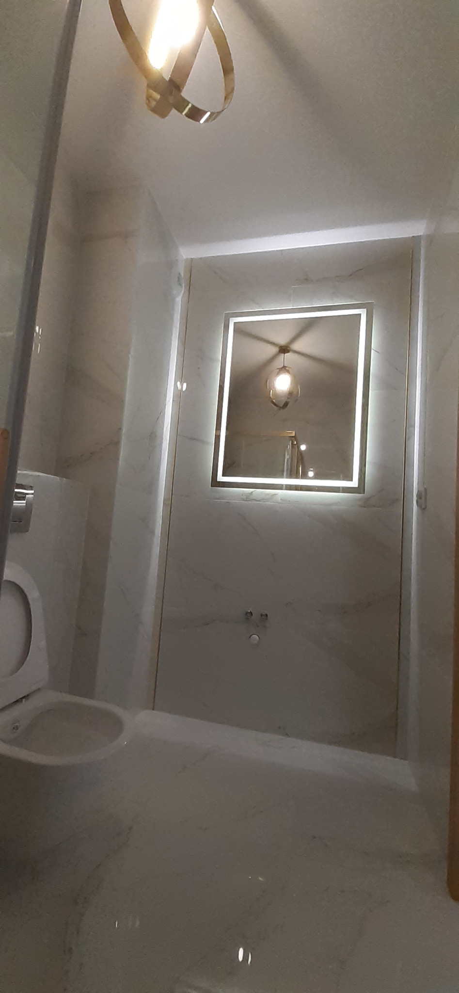 Wykończona łazienka w jasnych odcieniach z marmurowymi płytkami, podświetlanym lustrem w kształcie kwadratu i nowoczesną lampą sufitową.