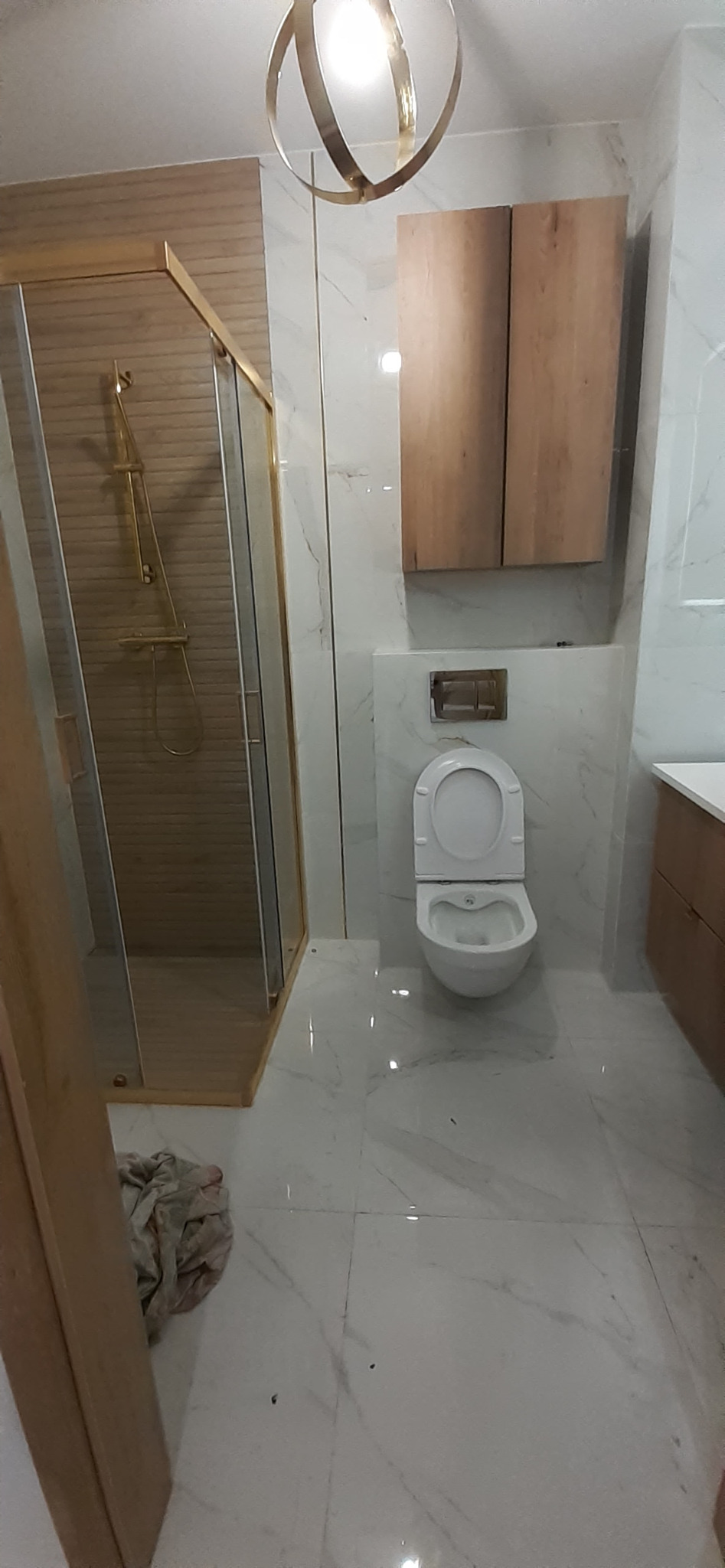 Wykończona łazienka w jasnych barwach z kabiną prysznicową ze złotymi ramami, podwieszaną toaletą i szafkami z naturalnego drewna oraz designerską lampą sufitową.