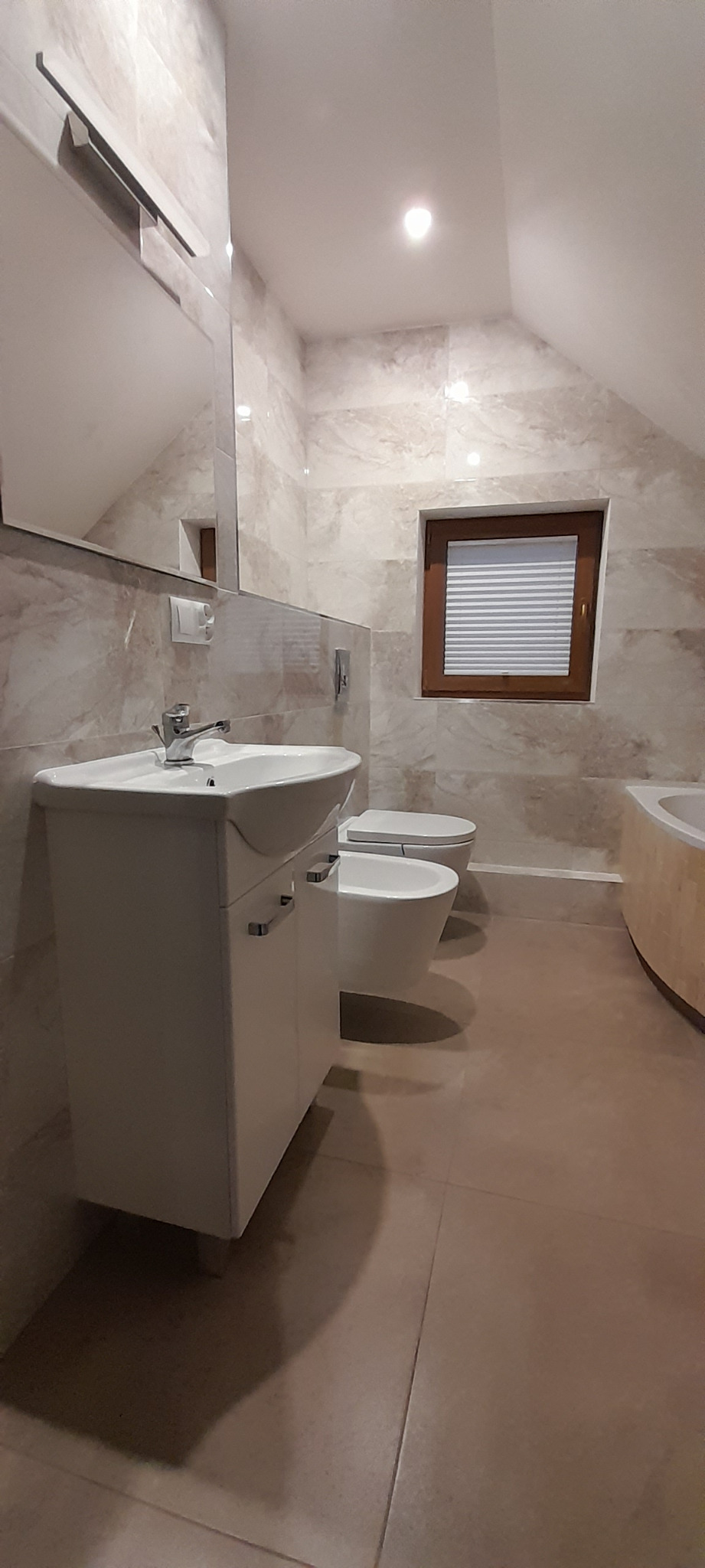 Wykończona łazienka na poddaszu ze skośnym sufitem, z umywalką z szafką w kolorze białym, toaletą, bidetem i oknem z drewnianą ramą oraz żaluzjami plisowanymi.