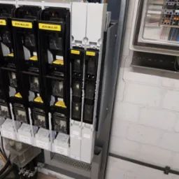 Rozdzielnica elektryczna z bezpiecznikami Jean Muller, widoczne kable i oznaczenia sekcji, zamontowana na ścianie z białej cegły.