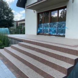 Wykończone schody wejściowe do domu z kamiennym dywanem w odcieniach brązu i beżu, z metalowymi listwami na krawędziach stopni, zabezpieczone folią drzwi balkonowe w tle.