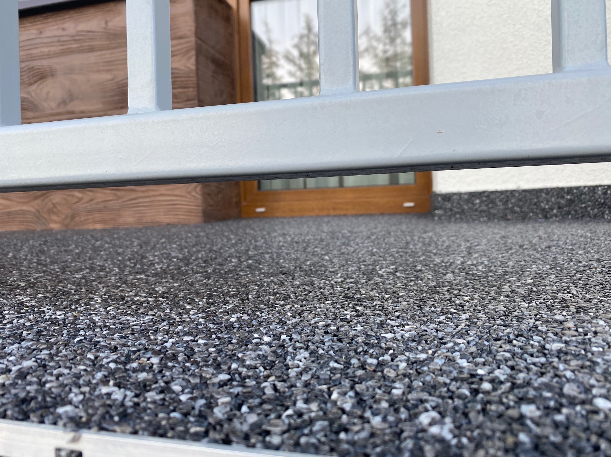 Szara posadzka z kamiennego dywanu na balkonie, widoczna przez metalową barierkę, w tle okno.