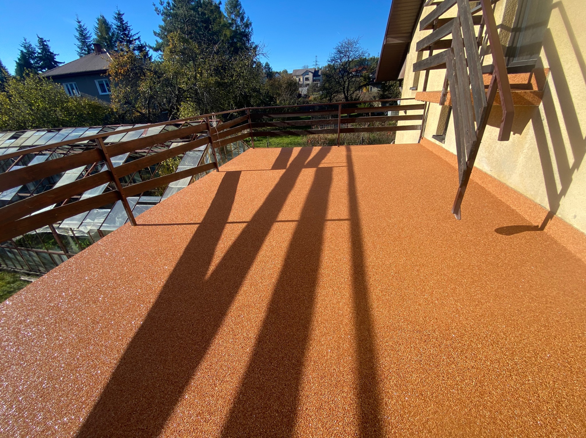 Balkon wykończony kamiennym dywanem w odcieniach brązu, z widocznymi cieniami barierki i schodów, na tle zieleni drzew i zabudowań.