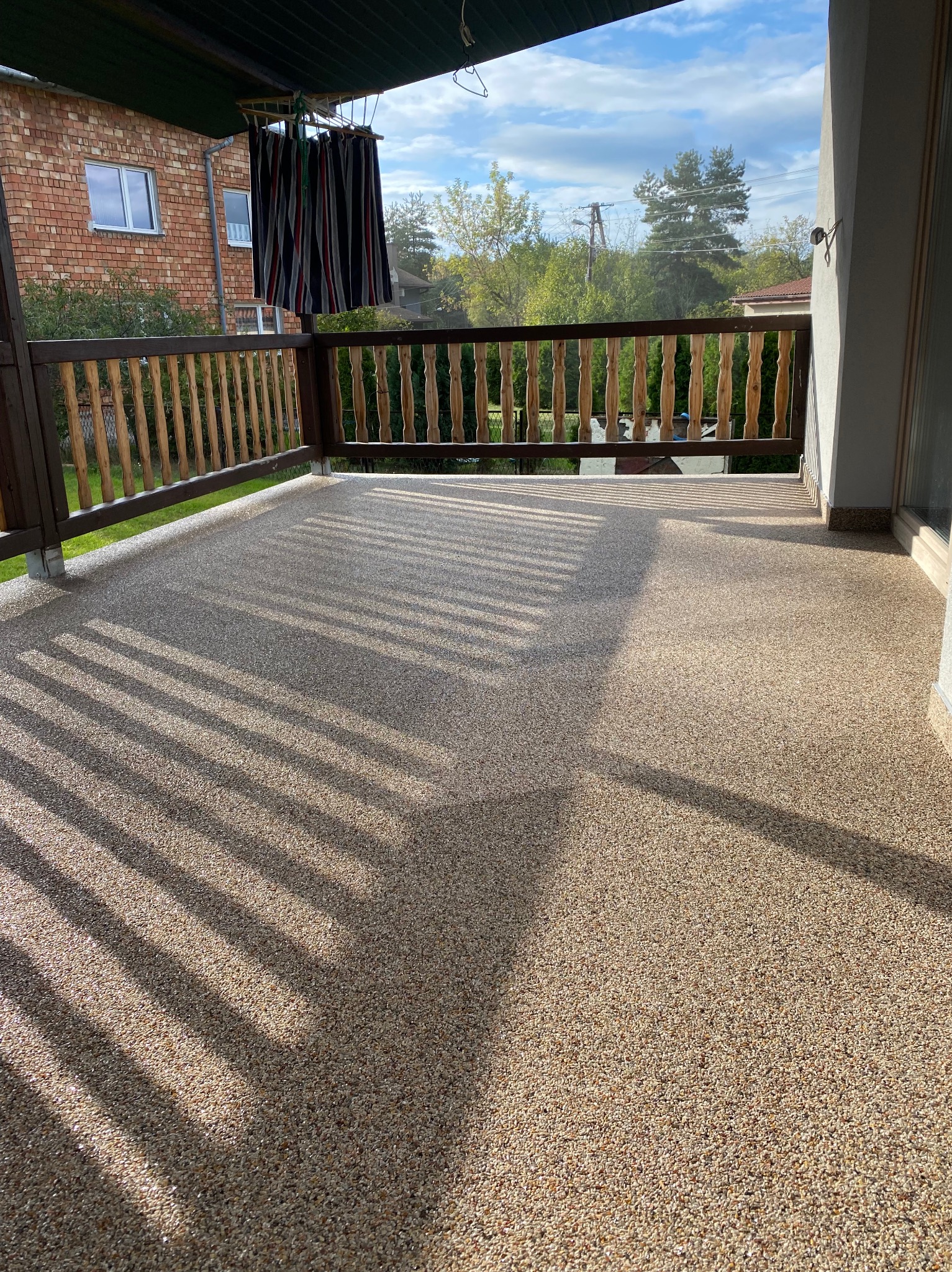 Beżowa, żwirowa posadzka na balkonie, z widocznymi cieniami barierki, w tle budynek mieszkalny i zieleń.