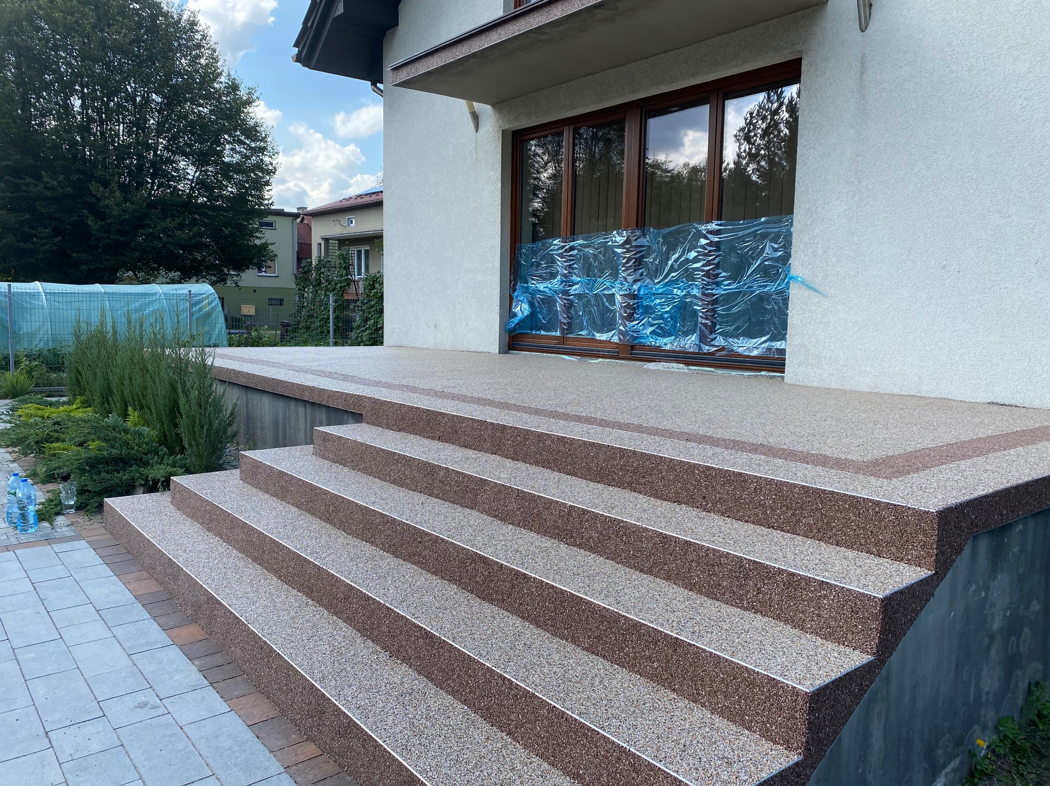 Wykończone schody wejściowe do domu z kamiennym dywanem w odcieniach brązu i beżu, z metalowymi listwami na krawędziach stopni, zabezpieczone folią drzwi balkonowe w tle.