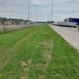 Shine-Usługi porządkowe - Długi pas zielonej trawy wzdłuż ogrodzenia i parkingu dla ciężarówek w pochmurny dzień, widoczne ślady kół na trawie.