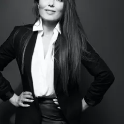 Czarno-białe zdjęcie portretowe kobiety w czarnej marynarce, białej koszuli i skórzanych spodniach, z rękami na biodrach, spoglądającej w górę.