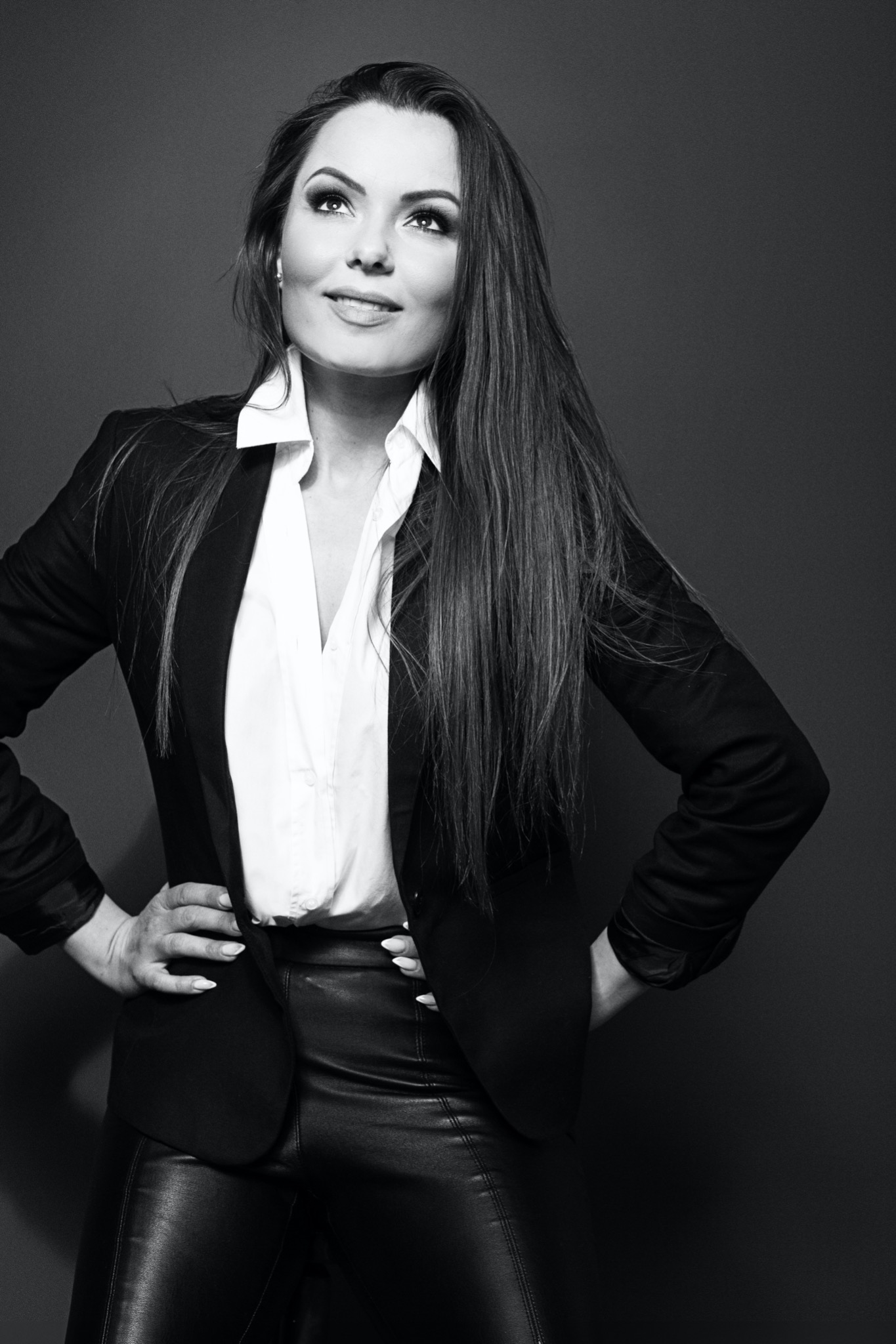 Czarno-białe zdjęcie portretowe kobiety w czarnej marynarce, białej koszuli i skórzanych spodniach, z rękami na biodrach, spoglądającej w górę.