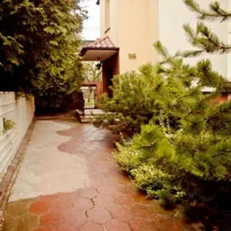 Wejście do budynku z numerem 16, wyłożone czerwoną kostką brukową, otoczone zielenią.