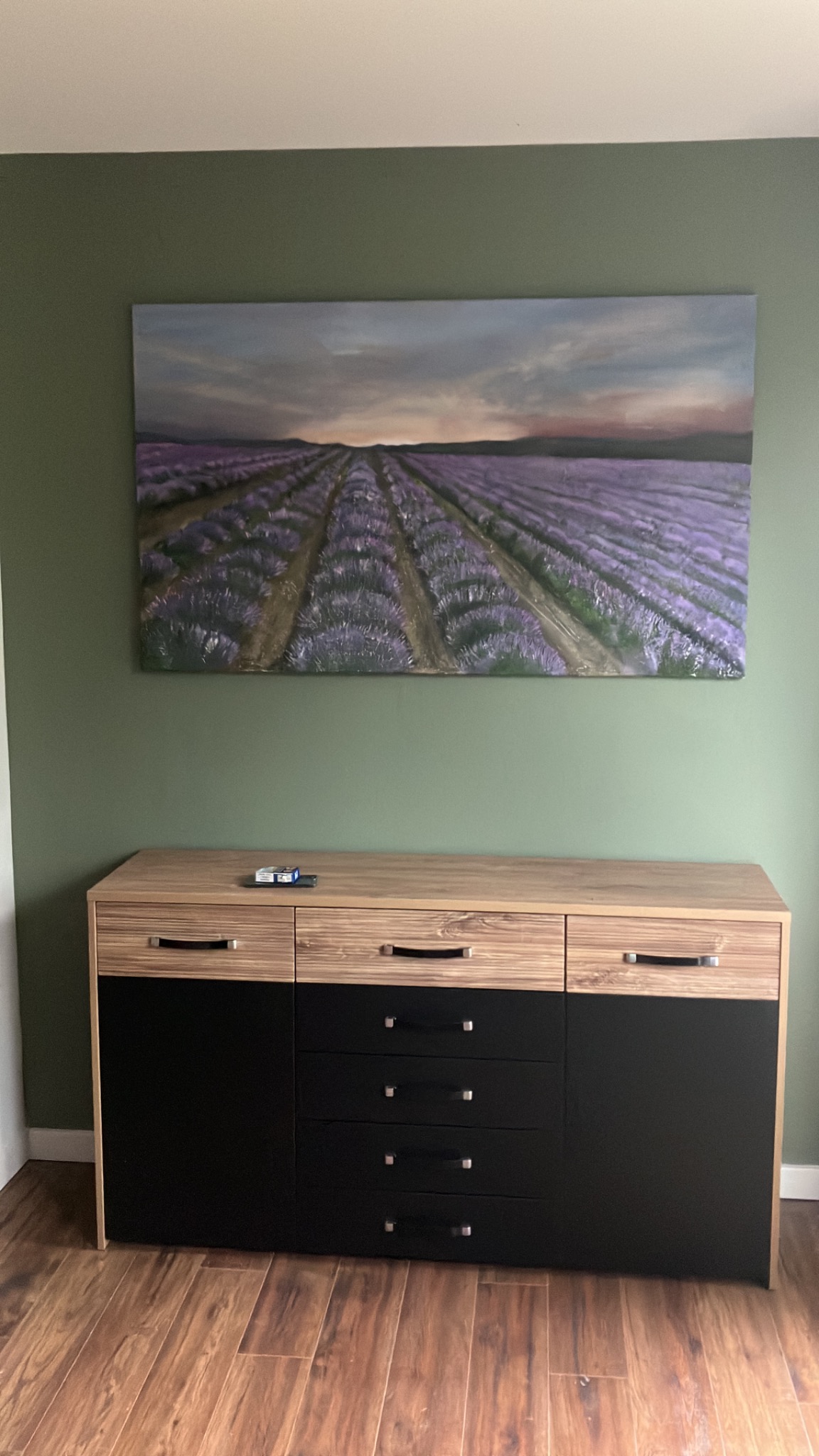 Komoda z jasnego drewna i czarnymi frontami szuflad, nad nią obraz z motywem lawendy na zielonej ścianie, wykończenie wnętrza.
