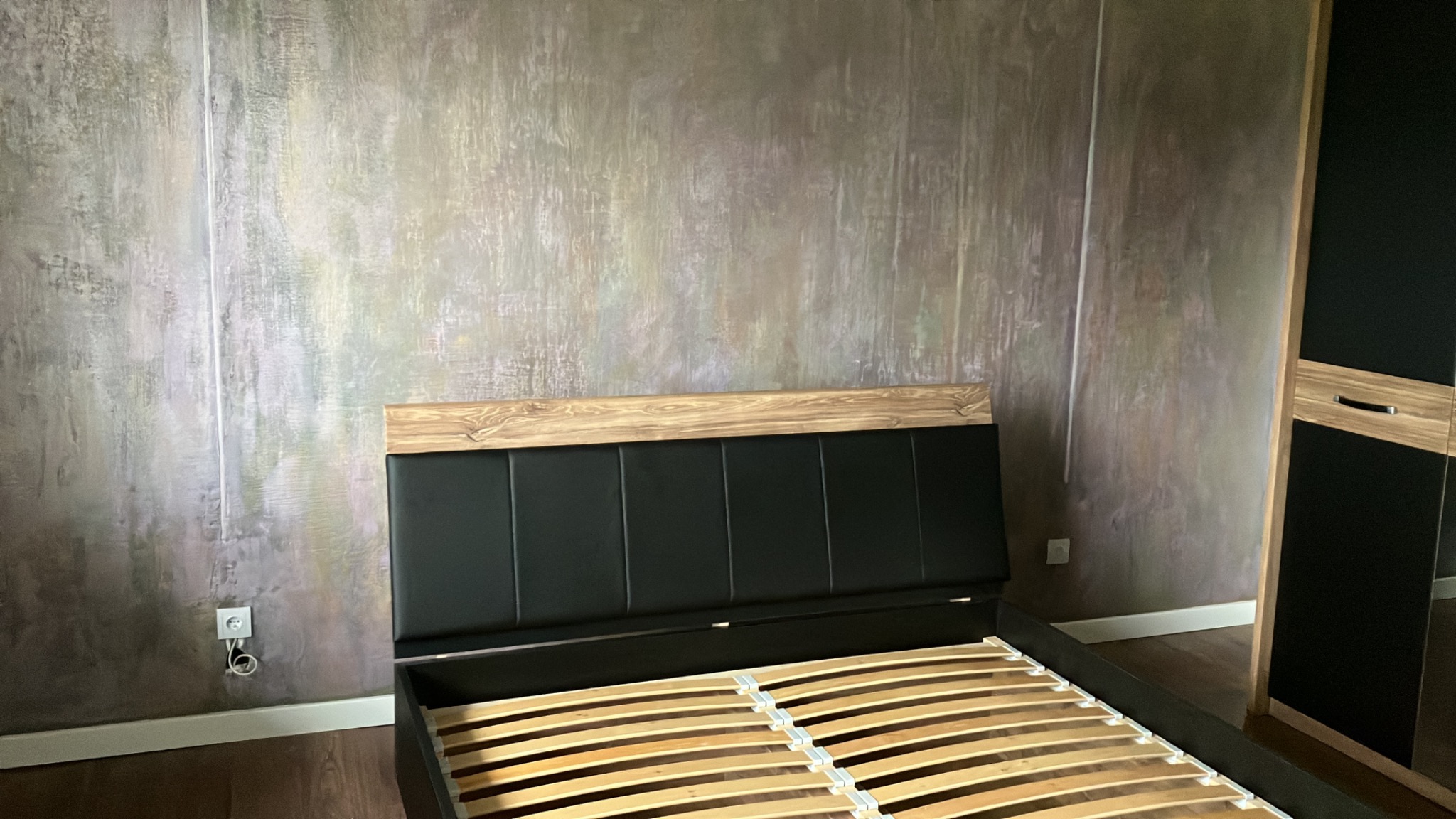 Ściana z efektem dekoracyjnym w Tarnowie, widoczna za ramą łóżka z drewnianym zagłówkiem i czarną tapicerką, obok szafa z czarnymi frontami i drewnianymi wstawkami.