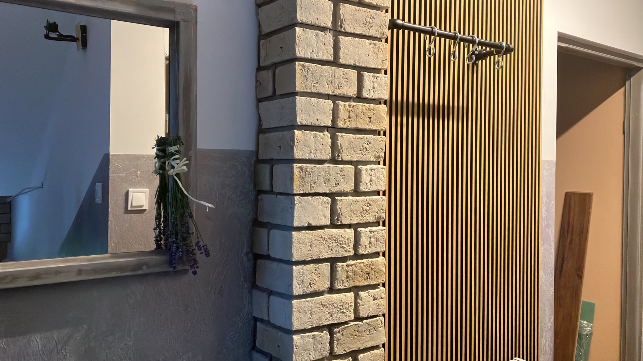 Fragment wykończonego wnętrza: dekoracyjna ściana z pionowych drewnianych listew z wieszakami, narożnik obłożony cegłą dekoracyjną, lustro w stylizowanej ramie i bukiet lawendy.