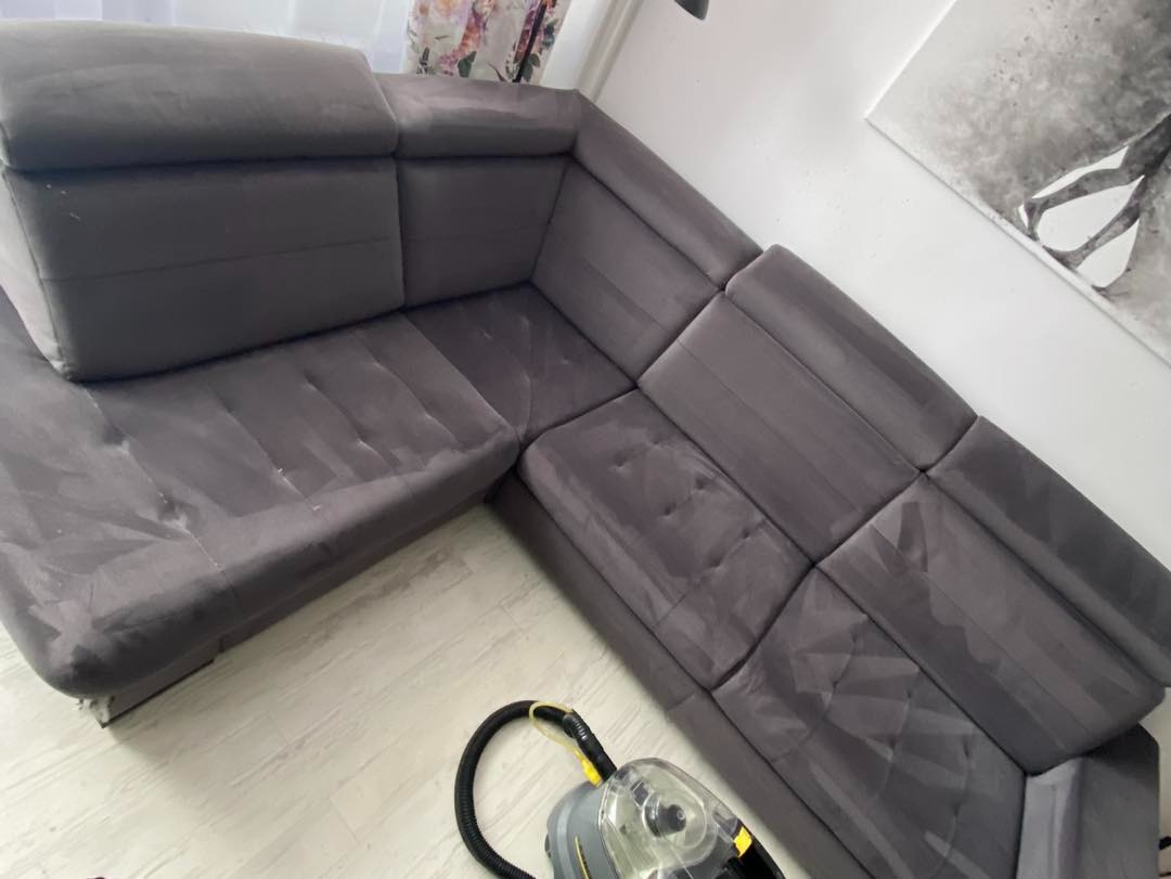 Szara narożna kanapa z widocznymi śladami czyszczenia i odkurzaczem piorącym na podłodze z paneli.