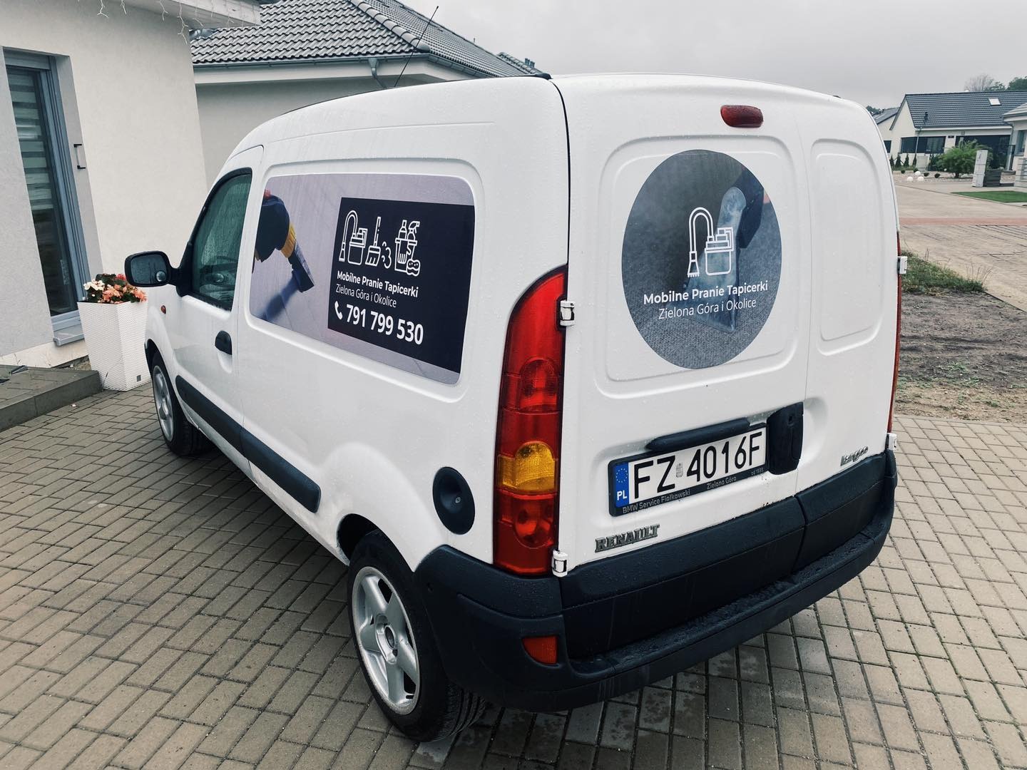 Biały samochód dostawczy z reklamą mobilnego prania tapicerki, zaparkowany na brukowanej nawierzchni przed domem.