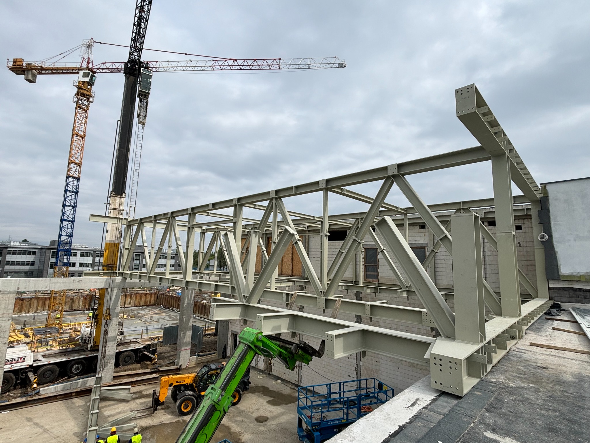 Montaż konstrukcji stalowej hali z użyciem dźwigów i podnośnika teleskopowego, widoczne elementy mocujące i pracownicy w żółtych kaskach.