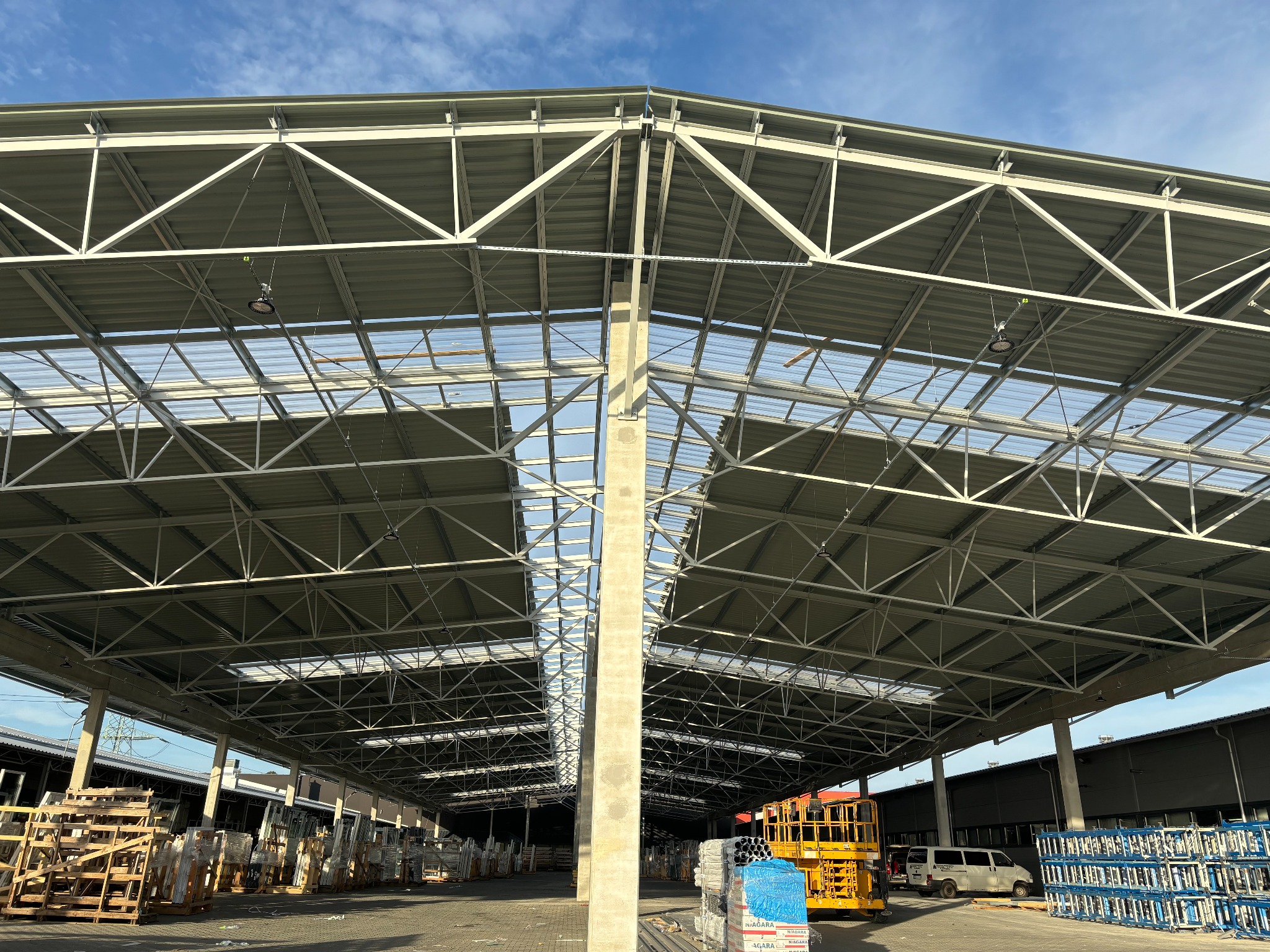 Wnętrze hali z widoczną konstrukcją stalową dachu, betonowymi słupami, paletami z materiałami budowlanymi i podnośnikiem koszowym.