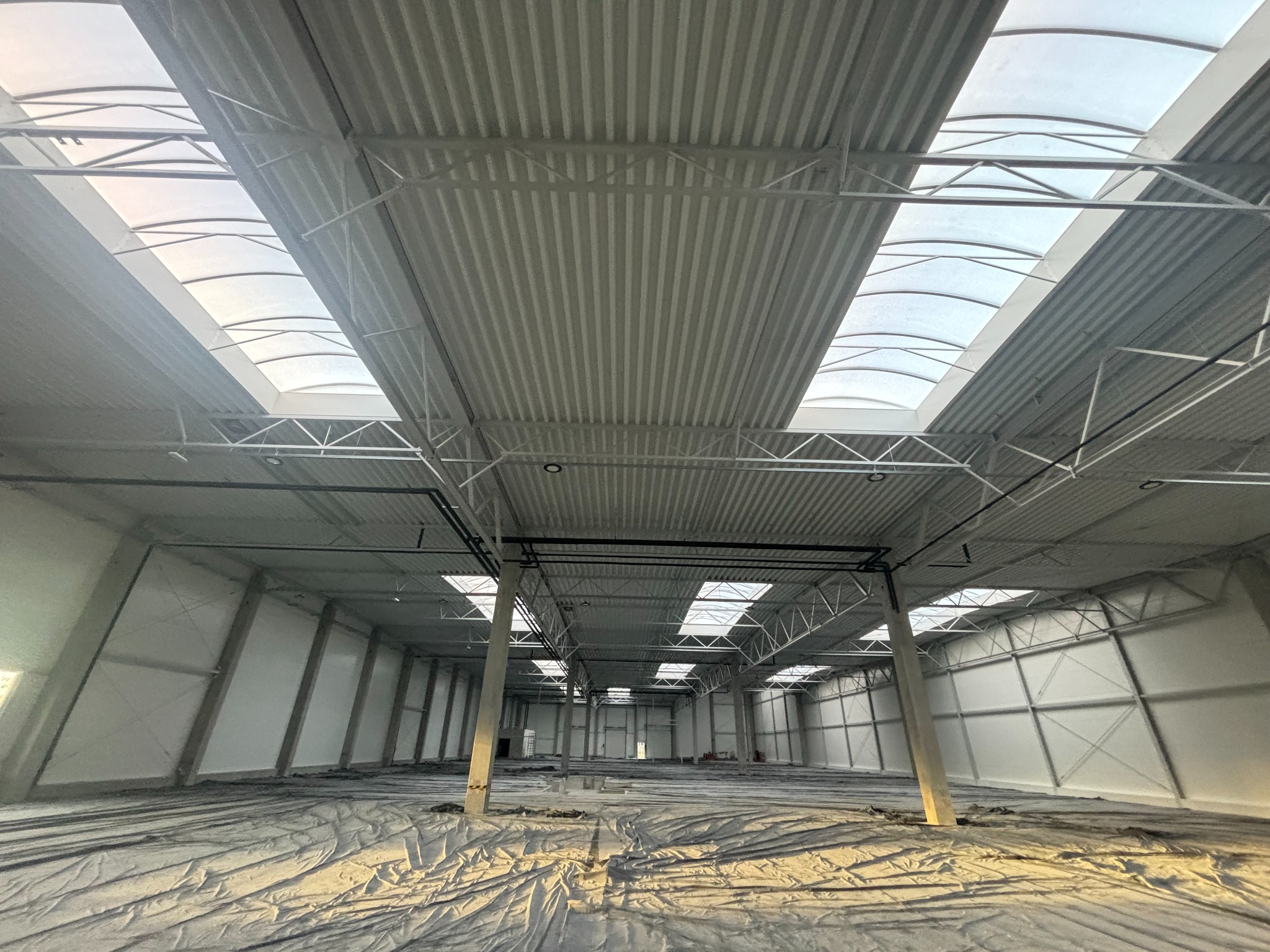Wnętrze nowo wybudowanej hali z widoczną konstrukcją stalową dachu i świetlikami dachowymi, podłoga zabezpieczona folią ochronną.