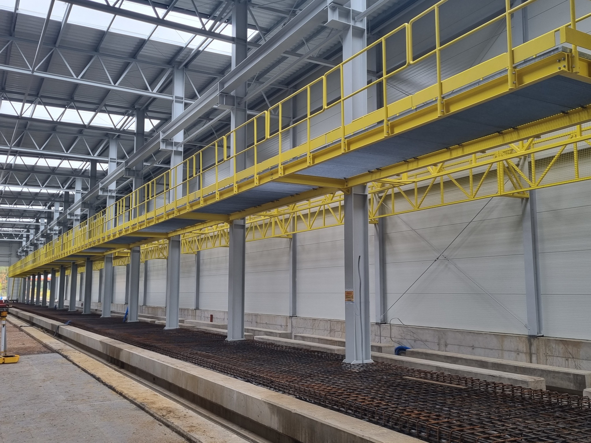 Wnętrze hali w trakcie budowy, widoczne stalowe konstrukcje nośne, żółte pomosty robocze i zbrojenie fundamentów.