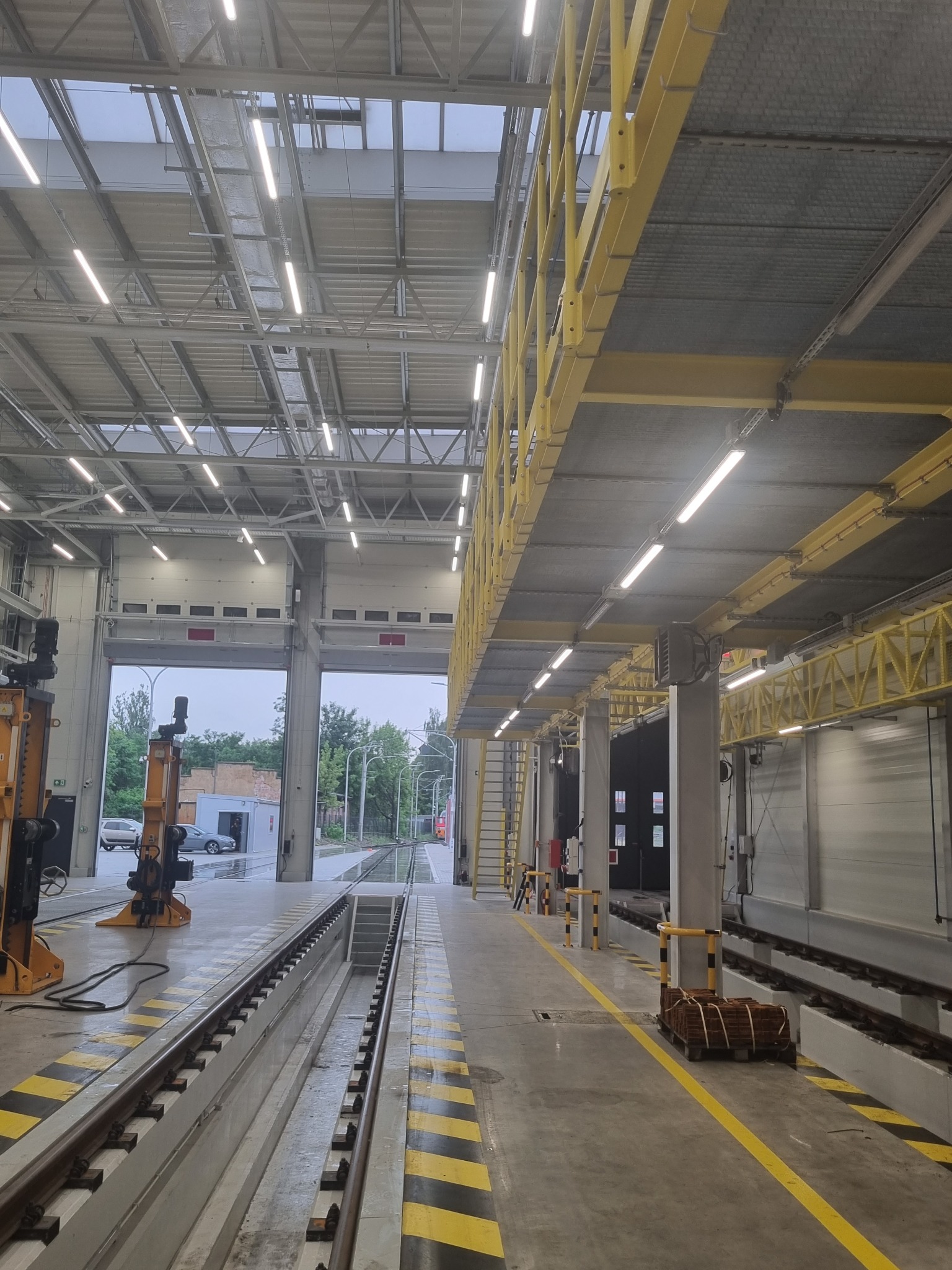 Wnętrze hali z torami kolejowymi i suwnicami, widok na otwarte bramy wjazdowe, oświetlenie jarzeniowe.