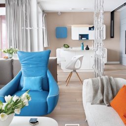 mDom Pracownia Projektowa - Jasne wnętrze mieszkania z akcentami błękitu: fotel z wysokim oparciem, modułowa biała kuchnia z błękitnymi elementami i dekoracyjna, przezroczysta ścianka działowa.