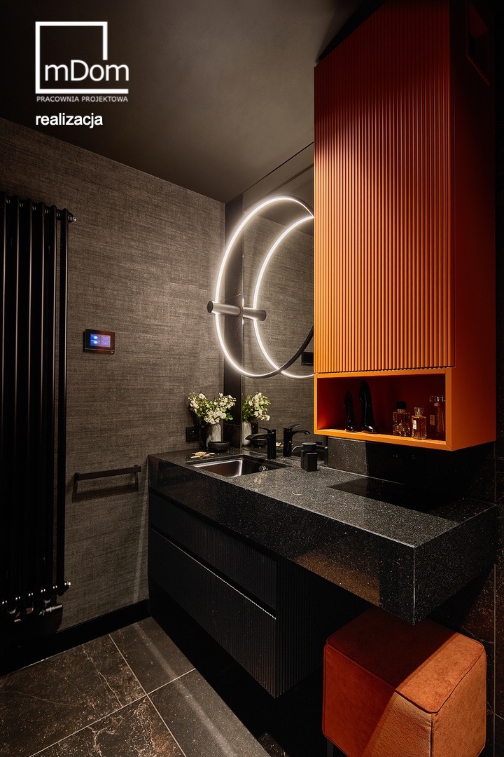 Nowoczesna łazienka z czarnym blatem z granitu, podświetlanym lustrem w kształcie okręgu, pomarańczową szafką z ryflowanymi frontami i czarnym grzejnikiem żeberkowym na tle szarej ściany z teksturą.