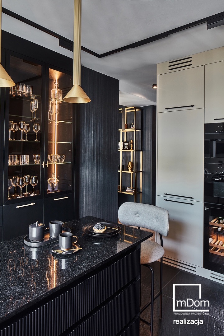 Wnętrze kuchni z wyspą wykończoną czarnym, żłobkowanym frontem, na której znajduje się czarna zastawa i deser z borówkami. W tle przeszklona szafka z podświetleniem i szklankami oraz regał...