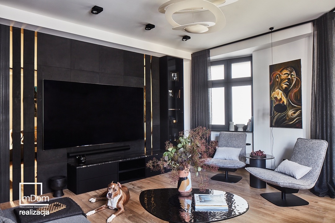 Elegancki salon z dużym telewizorem na ciemnej ścianie z złotymi akcentami, nowoczesnym oświetleniem i psem rasy pit bull leżącym na podłodze.