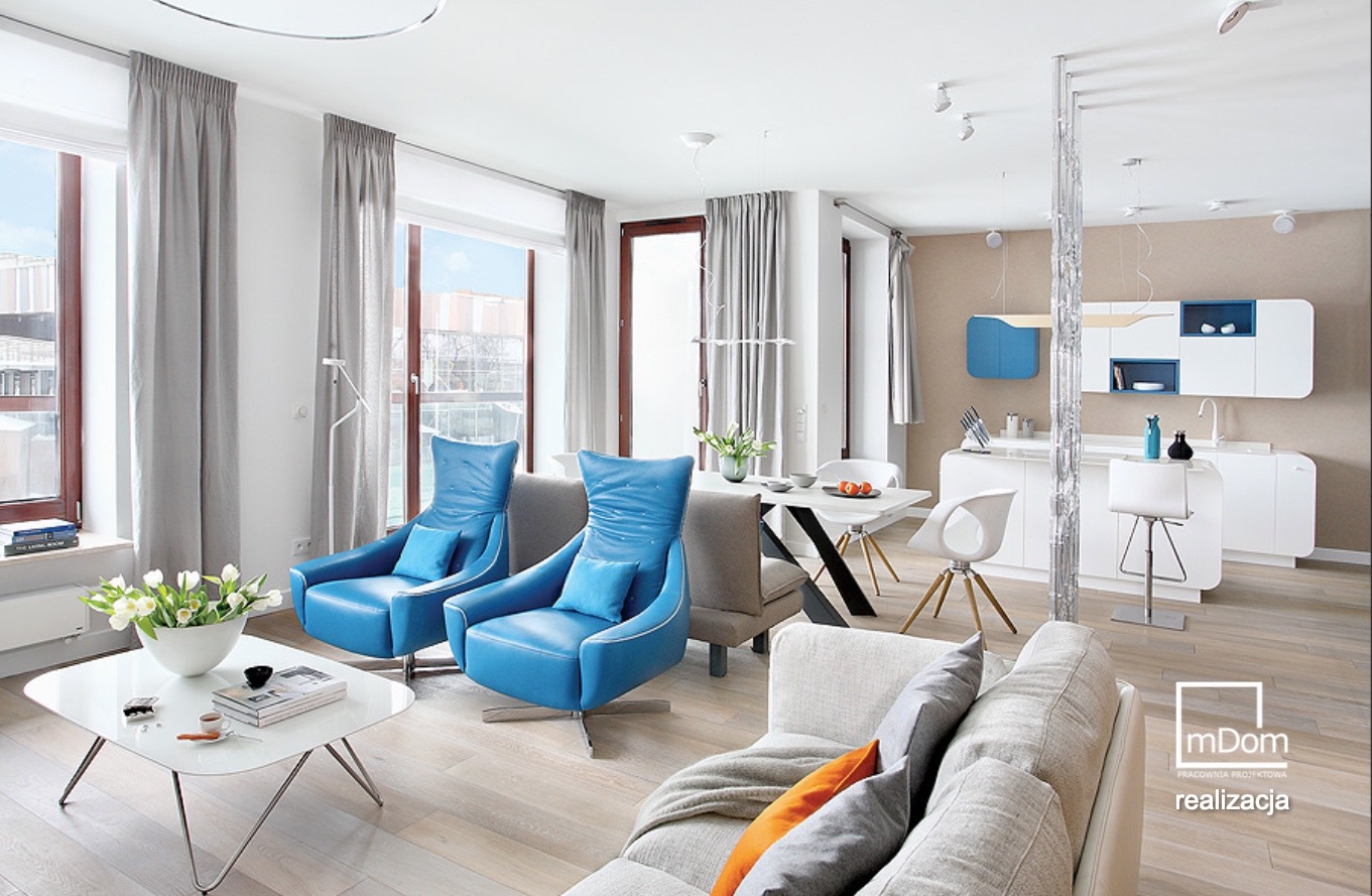 Jasne, przestronne wnętrze mieszkania z nowoczesnymi meblami: dwa błękitne fotele, biały stolik kawowy z tulipanami, stół jadalny z krzesłami i aneks kuchenny z białymi szafkami.
