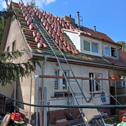 FAB DACH USŁUGI OGÓLNOBUDOWLANE - Dom w trakcie wymiany dachu: ułożone dachówki na połaci, rusztowania, widok z dołu, słoneczny dzień. Częściowo widoczne gałęzie drzew.