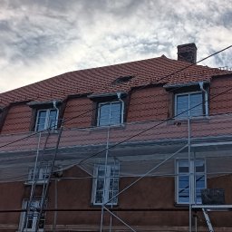 FAB DACH USŁUGI OGÓLNOBUDOWLANE - Ceglasty dach domu z lukarnami, częściowo osłonięty rusztowaniem. Widoczne rynny i komin. Tło stanowi pochmurne niebo i korony drzew.