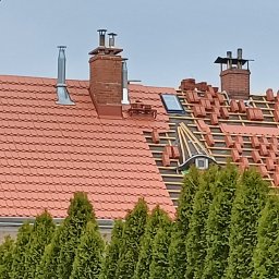 FAB DACH USŁUGI OGÓLNOBUDOWLANE - Częściowo pokryty dach z czerwonej dachówki, widoczne kominy i okno dachowe. Fragment dachu w trakcie krycia, z widoczną konstrukcją i ułożonymi dachówkami. W tle rząd zielonych drzew.