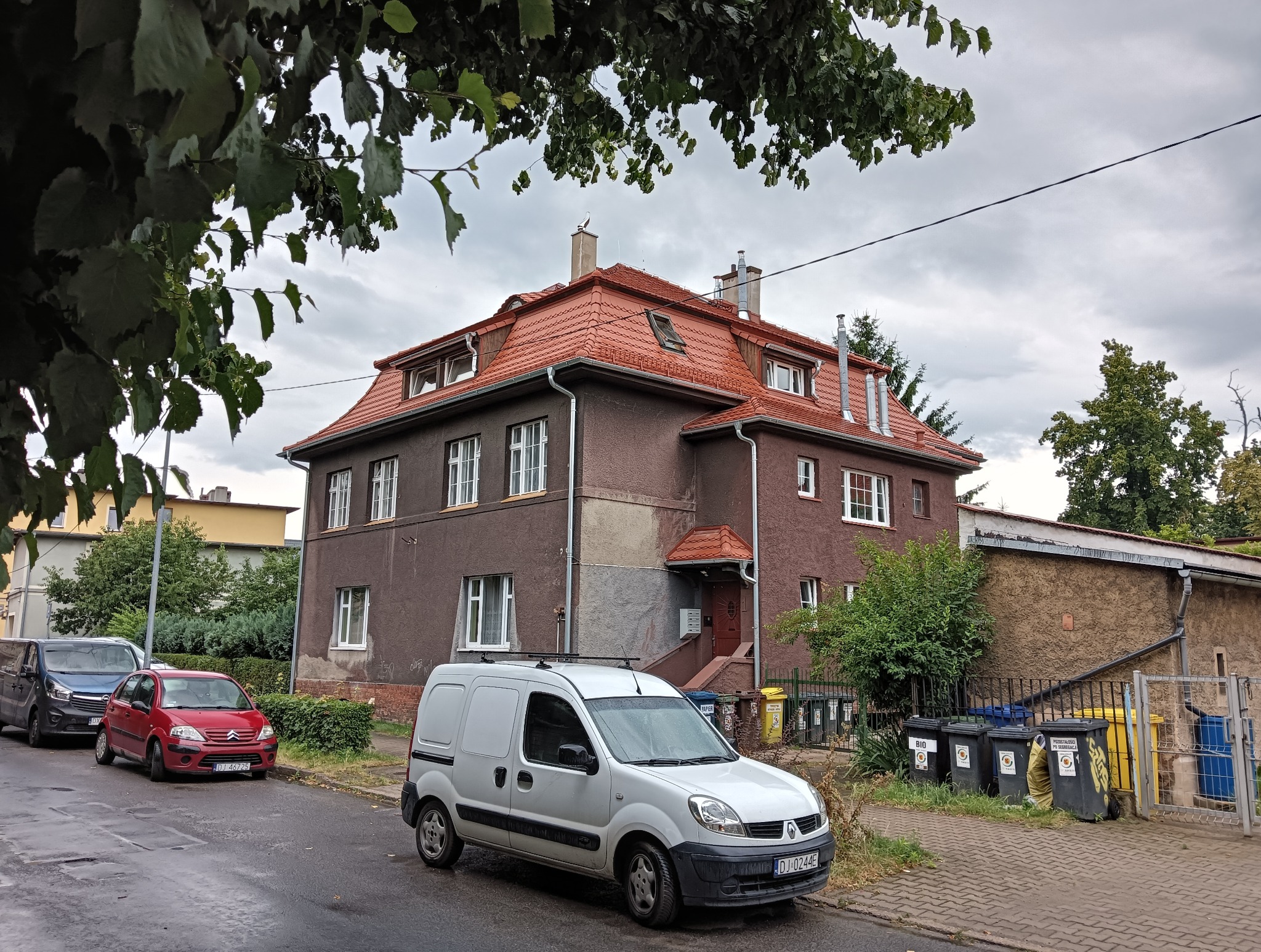 Dwupiętrowy dom z czerwonym dachem, widoczny z ulicy. Na dachu okna połaciowe i kominy. Przed budynkiem zaparkowany biały samochód dostawczy. Pochmurne niebo w tle.