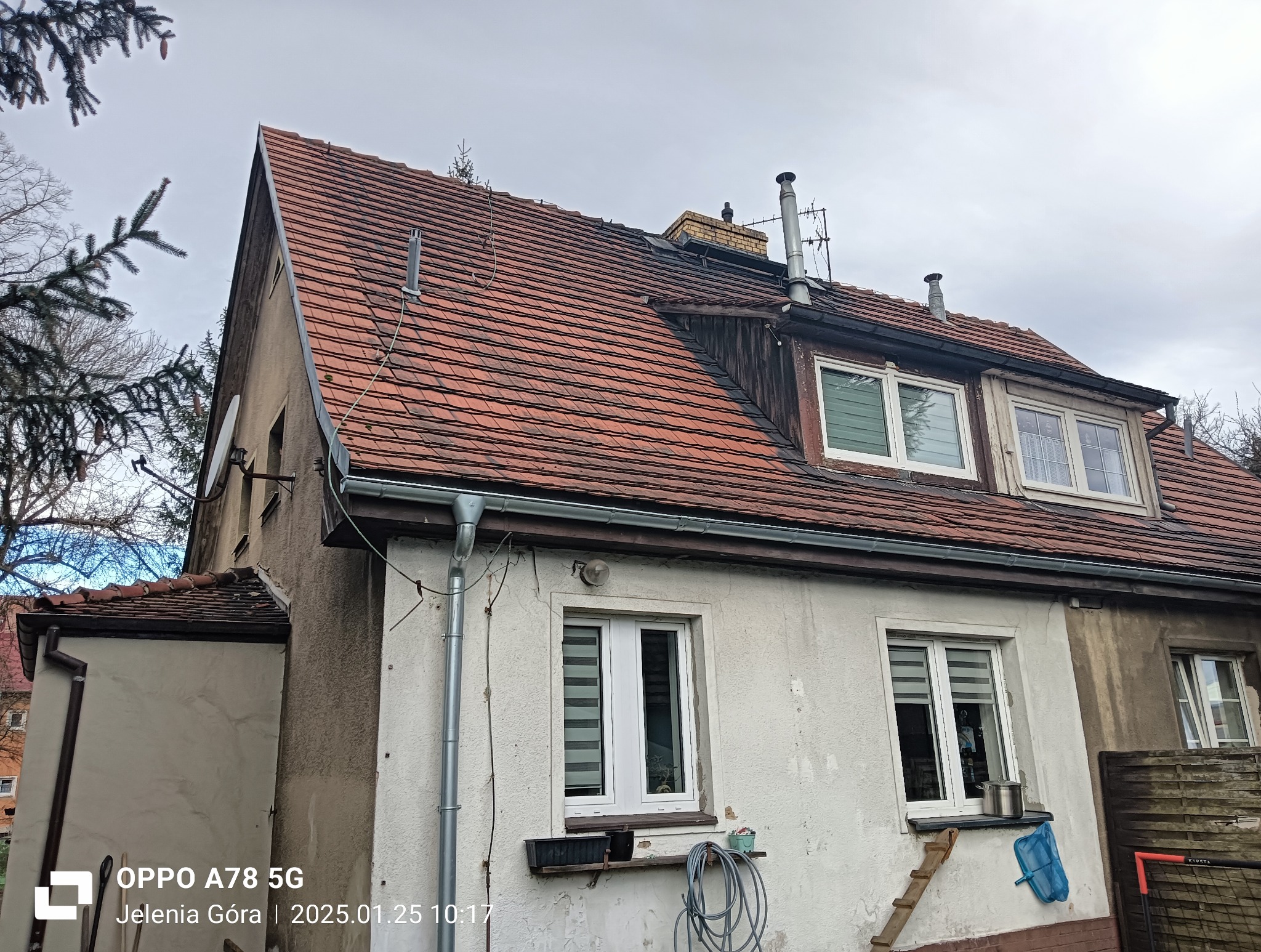 Dom z czerwoną dachówką widziany z dołu, z widocznymi oknami, rynnami i elementami elewacji. Dach pokryty dachówką ceramiczną, widoczne kominy i anteny.