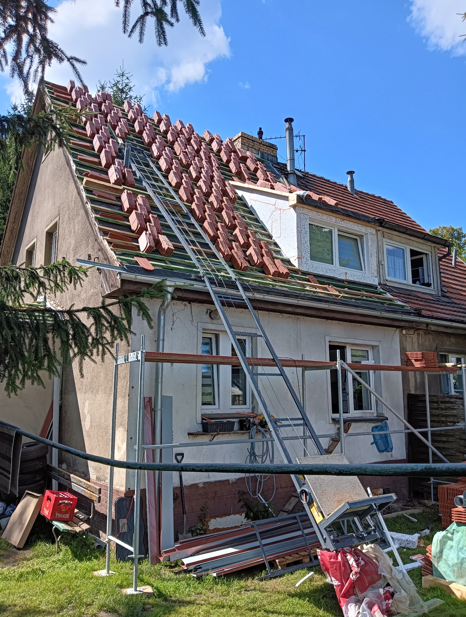 Dom w trakcie wymiany dachu: ułożone dachówki na połaci, rusztowania, widok z dołu, słoneczny dzień. Częściowo widoczne gałęzie drzew.
