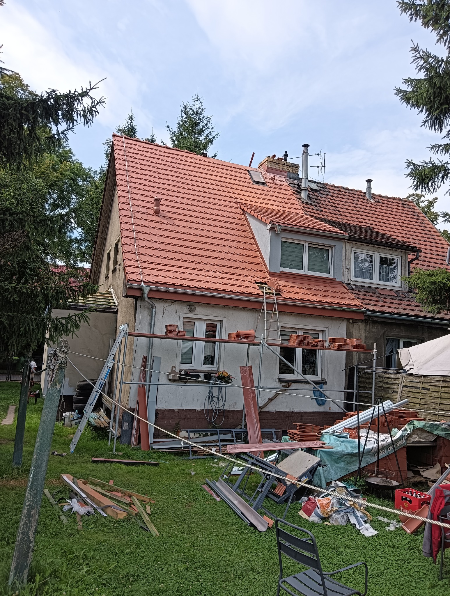 Dom z częściowo wymienionym, czerwonym dachem. Widoczne rusztowania, narzędzia i materiały budowlane. Prace dekarskie w toku, zieleń w tle.