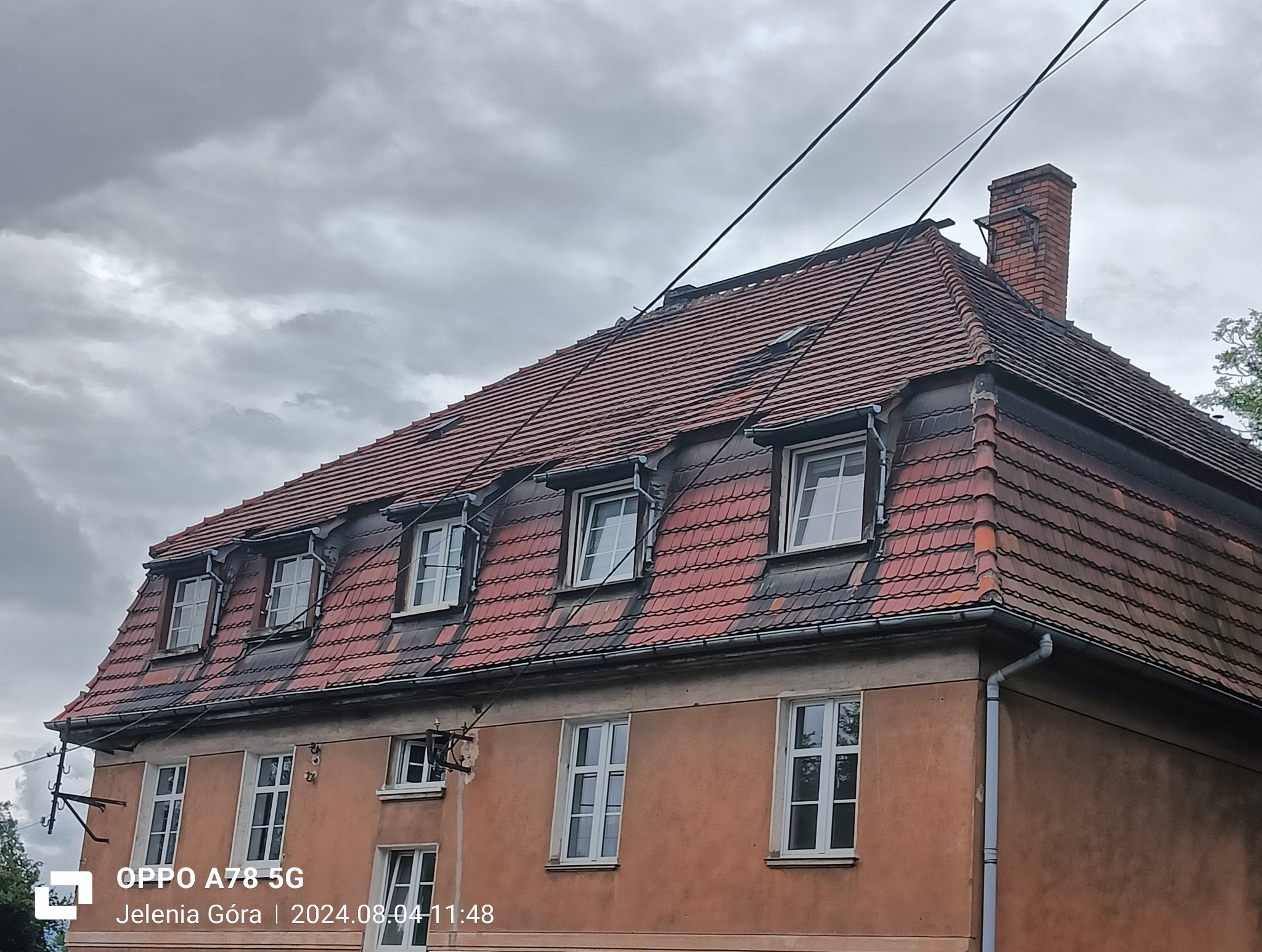 Ceglasty dach z lukarnami na kamienicy pod pochmurnym niebem. Widoczny komin i fragment elewacji. Dachówka karpiówka.