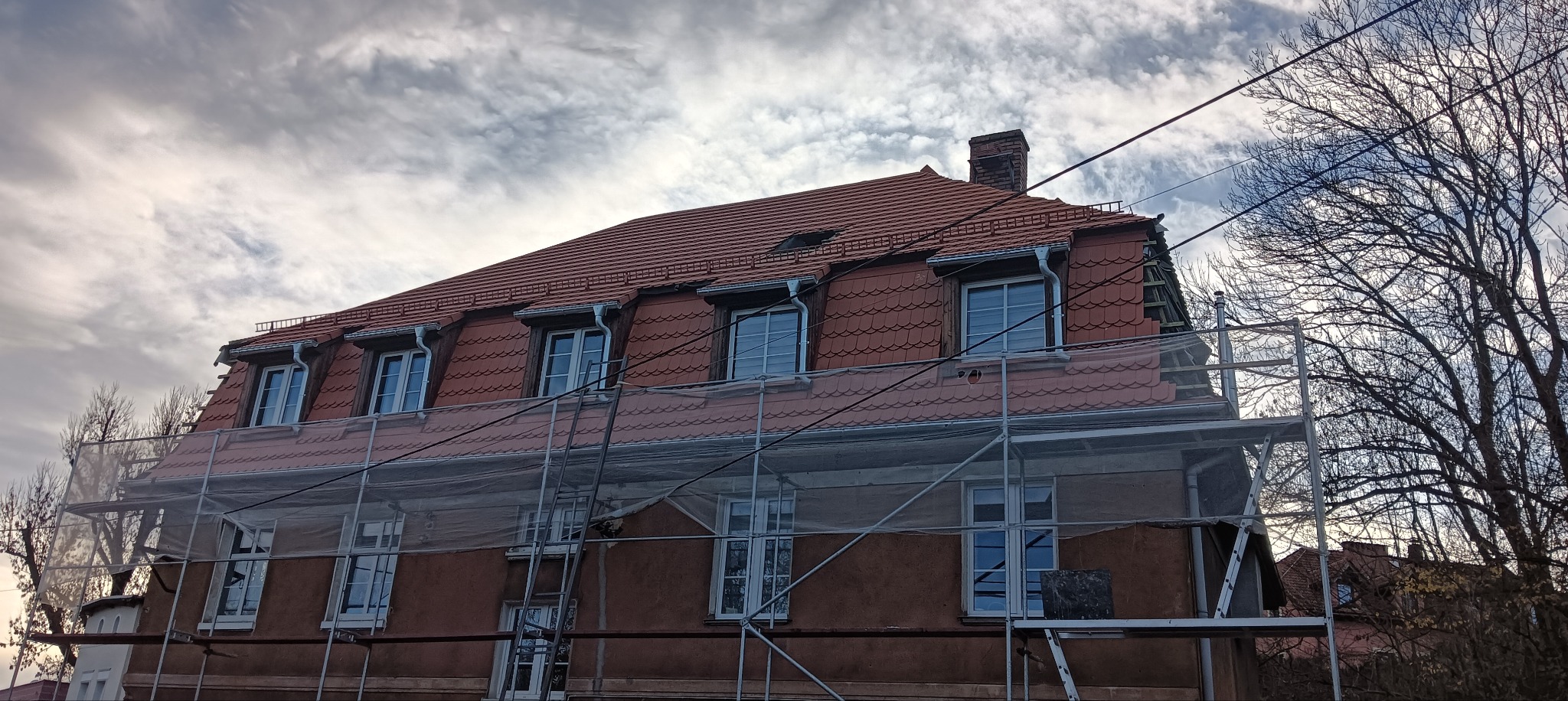Ceglasty dach domu z lukarnami, częściowo osłonięty rusztowaniem. Widoczne rynny i komin. Tło stanowi pochmurne niebo i korony drzew.