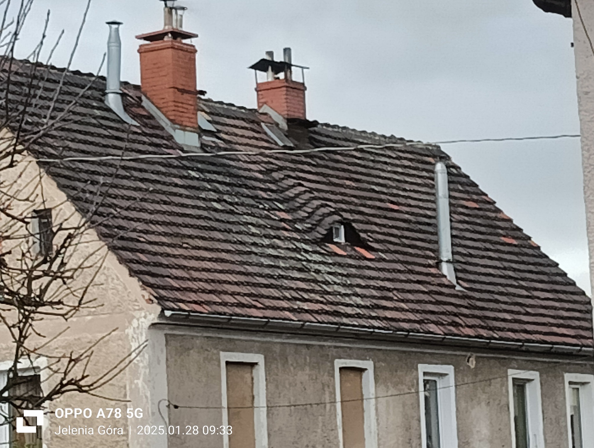 Stary dach z ceramicznej dachówki karpiówki, z widocznymi kominami i rurami wentylacyjnymi. Dachówka miejscami uszkodzona, z zapadnięciem w okolicy okna dachowego.