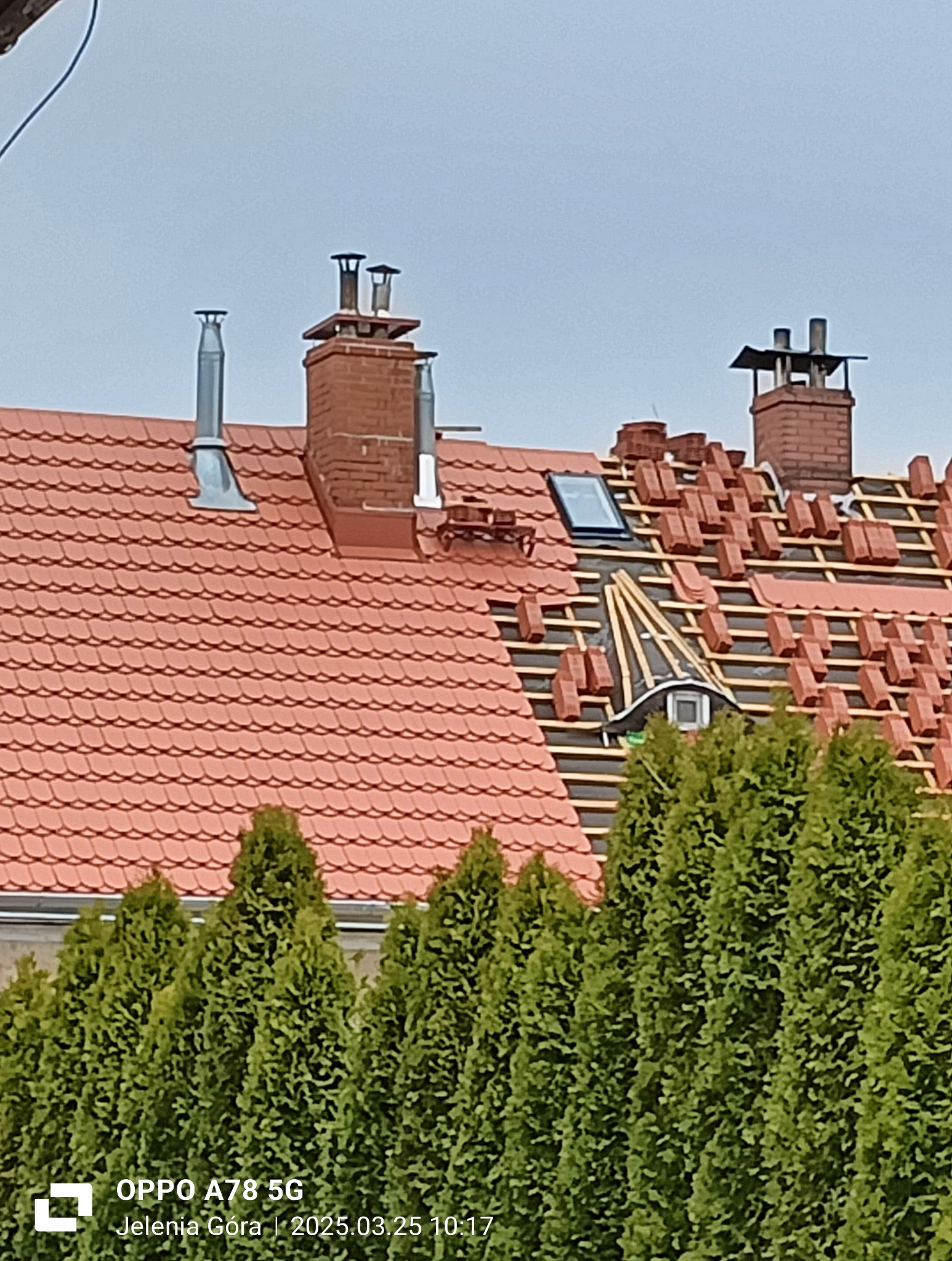 Częściowo pokryty dach z czerwonej dachówki, widoczne kominy i okno dachowe. Fragment dachu w trakcie krycia, z widoczną konstrukcją i ułożonymi dachówkami. W tle rząd zielonych drzew.