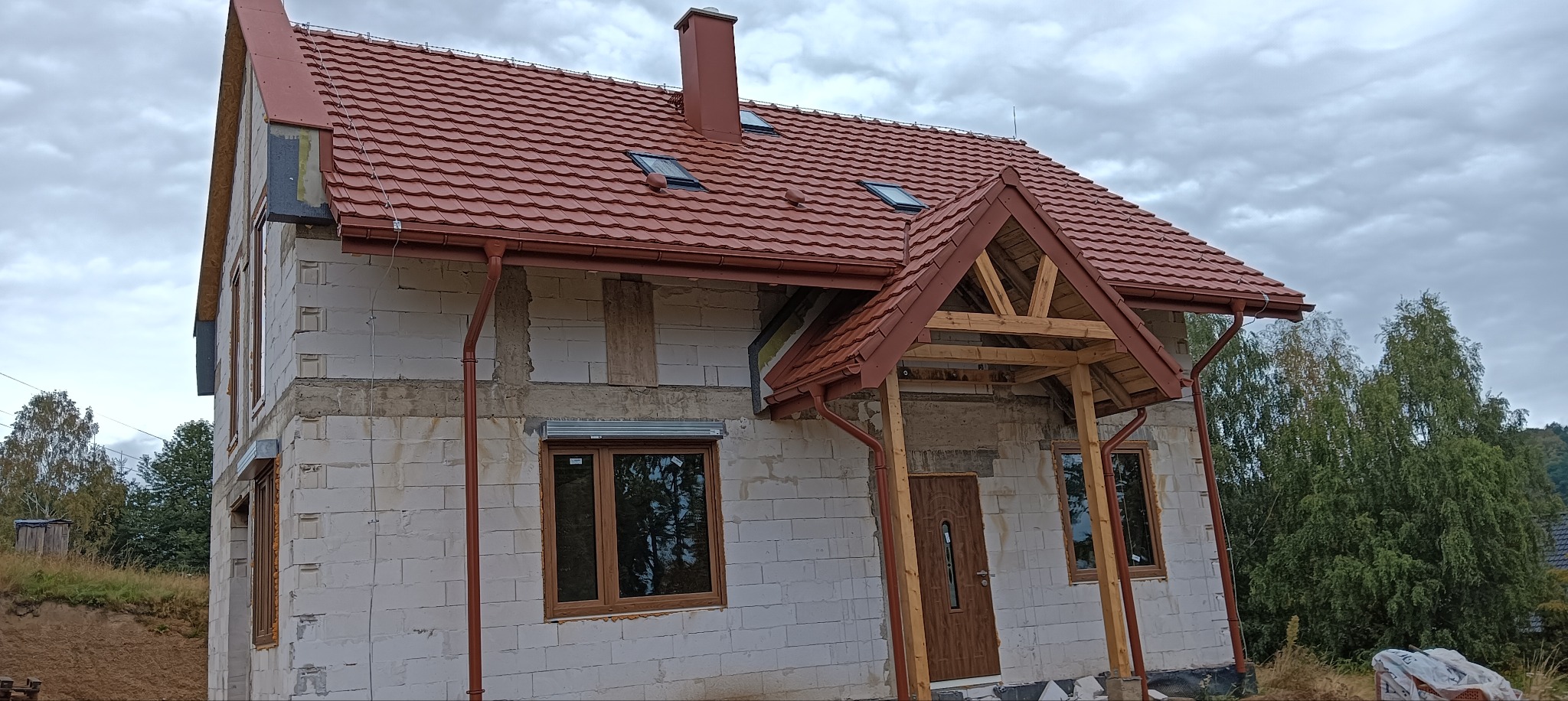 Dom w trakcie budowy z czerwoną dachówką, widoczne okna dachowe i orynnowanie. Częściowo wykończone wejście z drewnianą konstrukcją. Ściany z białego bloczka.