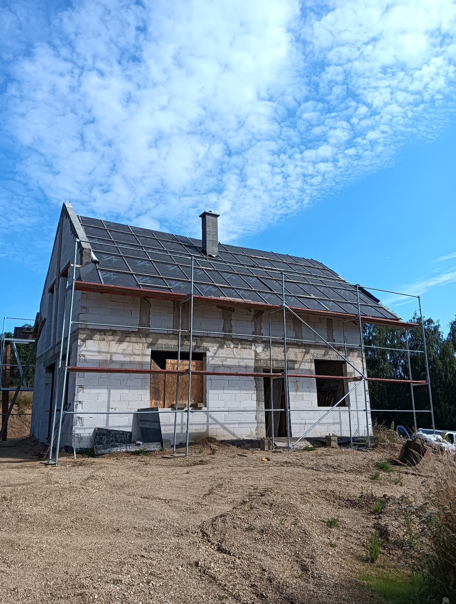 Dom w trakcie budowy z częściowo pokrytym dachem, otoczony rusztowaniami. Widoczne białe bloczki konstrukcyjne i czarna membrana dachowa na tle błękitnego nieba.